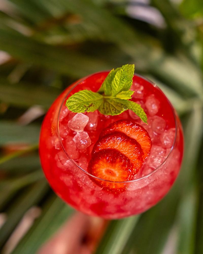 London Sour cocktail — vivid red strawberry sour over crushed ice with fresh strawberry slices and a mint sprig — set among tropical palm leaves